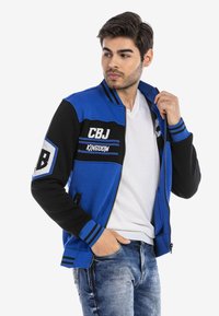 Blaue und schwarze Zip-Jacke mit gestreiften Bündchen und gestickten Patches, ausgestattet mit einem lässigen Schnitt und zwei vorderen Taschen.