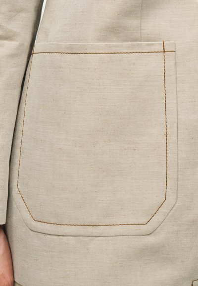 Μπεζ υφασμάτινη τσέπη με ορατές καφέ ραφές σε αντίστοιχο μπεζ ρούχο, τοποθετημένη κοντά στο ισχίο και το χέρι ενός ατόμου.