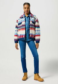 Chaqueta de manga larga con un patrón de diseños geométricos multicolores sobre un fondo crema, combinada con una camisa de mezclilla azul y jeans azules.