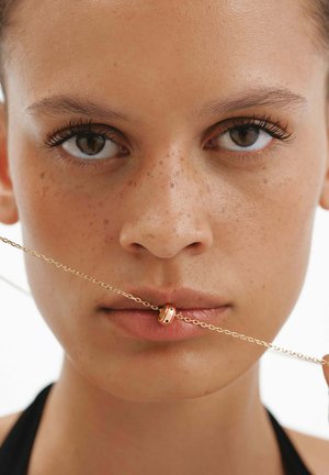 Collar de cadena de oro sostenido en los labios, con un colgante cilíndrico con acentos rojos y negros, sobre un fondo neutro.