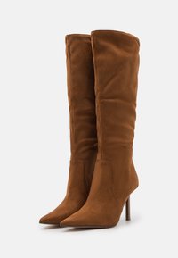 Par de botas altas de ante marrón con puntas afiladas y tacones de aguja delgados sobre un fondo blanco liso.