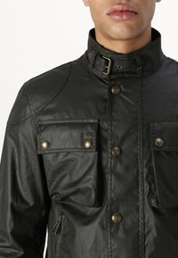 Black waterproof jacket with a high collar, two chest pockets, brass snap buttons, and a side zip pocket. Smooth texture with visible seams.