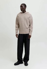 Felpa a maglia beige con una vestibilità rilassata, dotata di cappuccio e orlo a costine. Abbinata a pantaloni neri a righe verticali e scarpe nere.