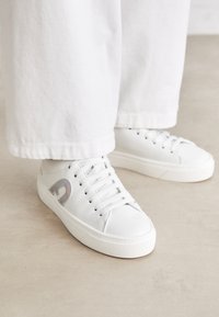 Sneaker bianco in pelle liscia con lacci bianchi, punta arrotondata e suola spessa bianca; presenta un accento riflettente sul lato.