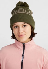 Cappello di lana verde a maglia con pom-pom in cima e testo marrone chiaro. Abbinato a una giacca in pile rosa con zip e logo nero.