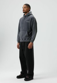 Sweat à capuche gris avec une poche kangourou, doté d'une capuche à cordon de serrage et d'un logo discret. Porté avec un pantalon noir et des chaussures noires avec des lacets roses.