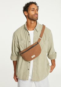 Sac en tissu marron en bandoulière avec fermeture éclair, forme arrondie et étiquette. Porté sur une chemise gris clair avec les manches retroussées.