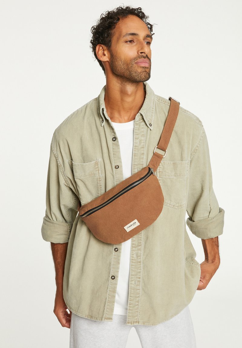 Sac en tissu marron en bandoulière avec fermeture éclair, forme arrondie et étiquette. Porté sur une chemise gris clair avec les manches retroussées.