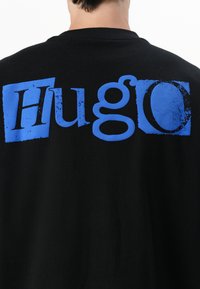 Camiseta de algodón negra con un diseño gráfico desgastado en azul que dice "HuGo" en la parte posterior. Diseño clásico de cuello redondo con mangas cortas.
