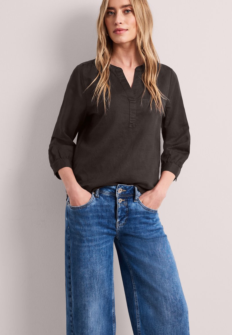 Femme aux longs cheveux blonds portant un chemisier noir à manches longues et un jean bleu taille haute, les mains dans les poches, sur un fond uni.