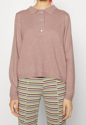 Femme portant un pull en maille rose poudré avec trois boutons décoratifs et un pantalon côtelé rayé multicolore, se tenant devant un fond uni.