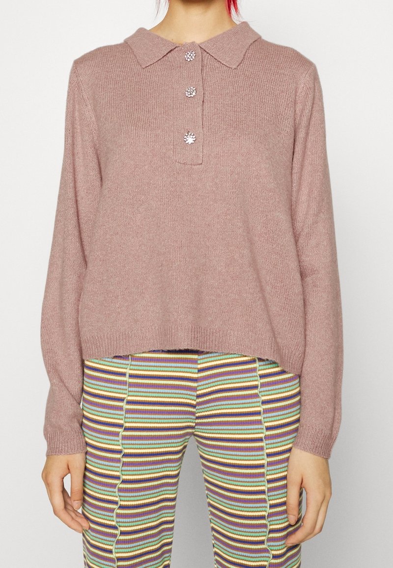 Femme portant un pull en maille rose poudré avec trois boutons décoratifs et un pantalon côtelé rayé multicolore, se tenant devant un fond uni.