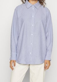 Raidallinen nappikauluspaita, jossa on vaaleansininen ja valkoinen värimaailma. Paidassa on kaulus, pitkät hihat ja rento leikkaus. Kangas näyttää sileältä.