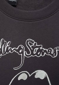 Svart sweatshirt med ribbad halsringning, med vit grafisk text "Rolling Stones" och ikonisk läpdesign i silver. Mjuk, texturerad tyg.