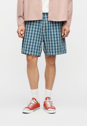 Férfi, aki kék-fehér kockás rövidnadrágot, rózsaszín, gombos inget, fehér zoknit és piros Converse tornacipőt visel, fehér háttér előtt áll.