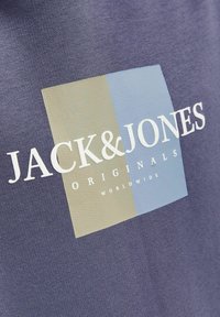 Getextureerde stof in marineblauw met een vierkant logo in gedempt groen en lichtblauw. Bevat witte merktekst: "JACK & JONES ORIGINALS WORLDWIDE."