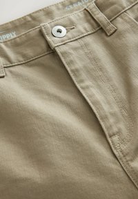Pantalons ajustés beiges en coton. Dotés d'un bouton en métal et d'une fermeture éclair, avec des coutures visibles et une finition en tissu texturé.