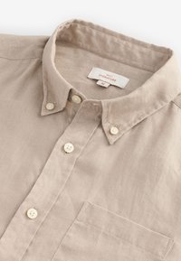 Hellbeiges Leinenhemd mit Button-down-Kragen, vier Knöpfen vorne und einer Brusttasche. Das Etikett trägt die Aufschrift "NEXT SIGNATURE M."