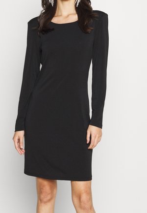 Woman wearing a black knee-length dress with long sleeves and padded shoulders, paired with dangling crystal earrings.