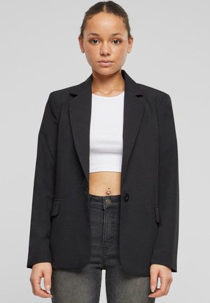 Jeune femme portant un blazer noir par-dessus un top court blanc et un jean taille haute gris foncé, debout devant un fond gris uni.