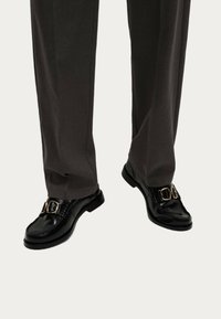 Zwarte leren loafers met gouden gespaccenten, gecombineerd met donkergrijze, op maat gemaakte broeken. De nadruk ligt op het slanke ontwerp en de glanzende afwerking.