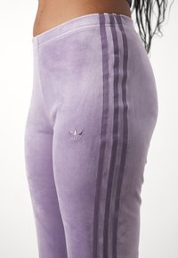 Nära bild av en person som bär högmidjade ljuslila velourbyxor med tre vertikala ränder och en liten logotyp på låret.