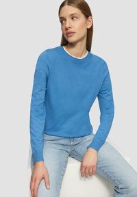 Blauer Strickpullover mit Rundhalsausschnitt und langen Ärmeln, getragen über hellblauen Jeans, sitzt auf einer weißen Fläche.