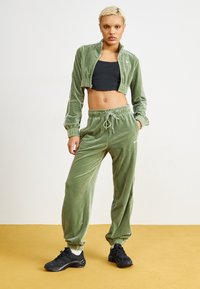 Velour-matchande set i ljusgrön; kort zip-jacka med elastiska ärmar, joggerbyxor med dragsko i midjan och elastiska anklar. Svarta sneakers.