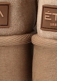 Zapatillas de ante color tostado con un exterior texturizado, forma redondeada y un prominente parche de logo marrón con letras en relieve en el lateral.