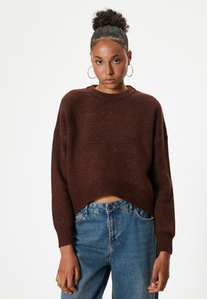 Brown knitted sweater with a cropped design, round neckline, and ribbed cuffs. Worn over blue denim jeans, showcasing a relaxed fit.