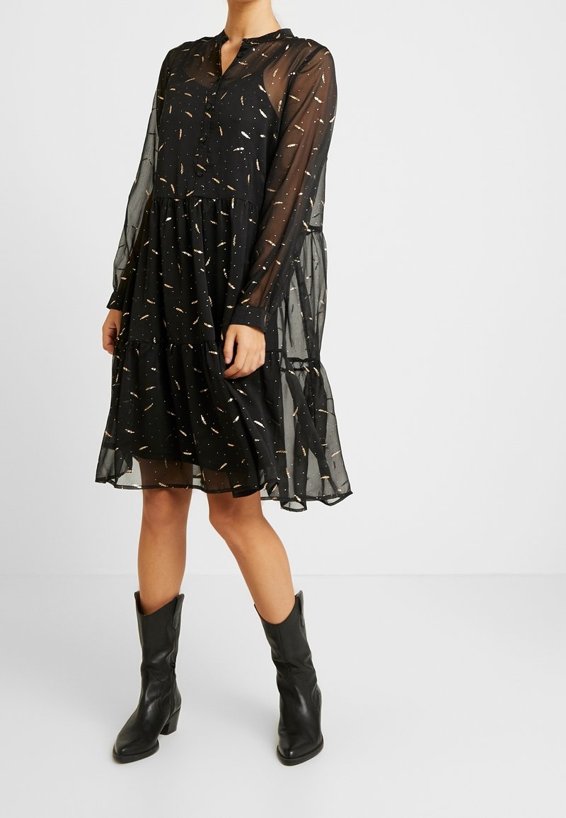 Svart, genomskinlig klänning med långa ärmar, knäppt framtill och med flätad kjol, prydd med guldprickar. Matchas med svarta, klackiga cowboystövlar.