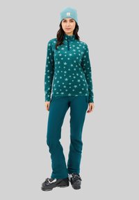 Pullover in fleece color turchese con motivo a stelle, colletto a mezza zip; abbinato a pantaloni turchesi e scarponi da sci neri. Berretto lavorato a maglia color blu chiaro sulla testa.
