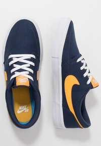 Merkkikenkien sininen kangaskenkä valkoisilla nauhoilla ja kumipohjilla. Sivussa on oranssi Nike-logo ja sininen pohjallinen.