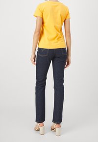 T-shirt gialla in cotone a maniche corte abbinata a jeans in denim blu scuro. Il modello indossa sandali beige con tacco. Design semplice, senza motivi visibili.