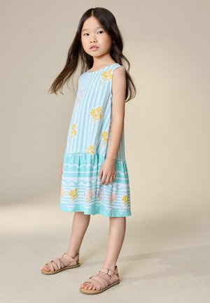 Jauna meitene stāv, valkājot bezroku tirkīzzilu kleitu ar dzelteniem ziedu rakstiem un gaiši rozā sandales, neitrāla fona.