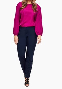 Blouse en satin fuchsia avec manches longues bouffantes, associée à un pantalon bleu marine slim. Des talons noirs à brides complètent la tenue.
