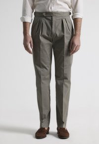 Pantalones de tejido khaki con pliegues y un corte ajustado, que cuentan con una cintura con botones, una textura suave y piernas rectas. Acentuados con zapatos de ante marrón.