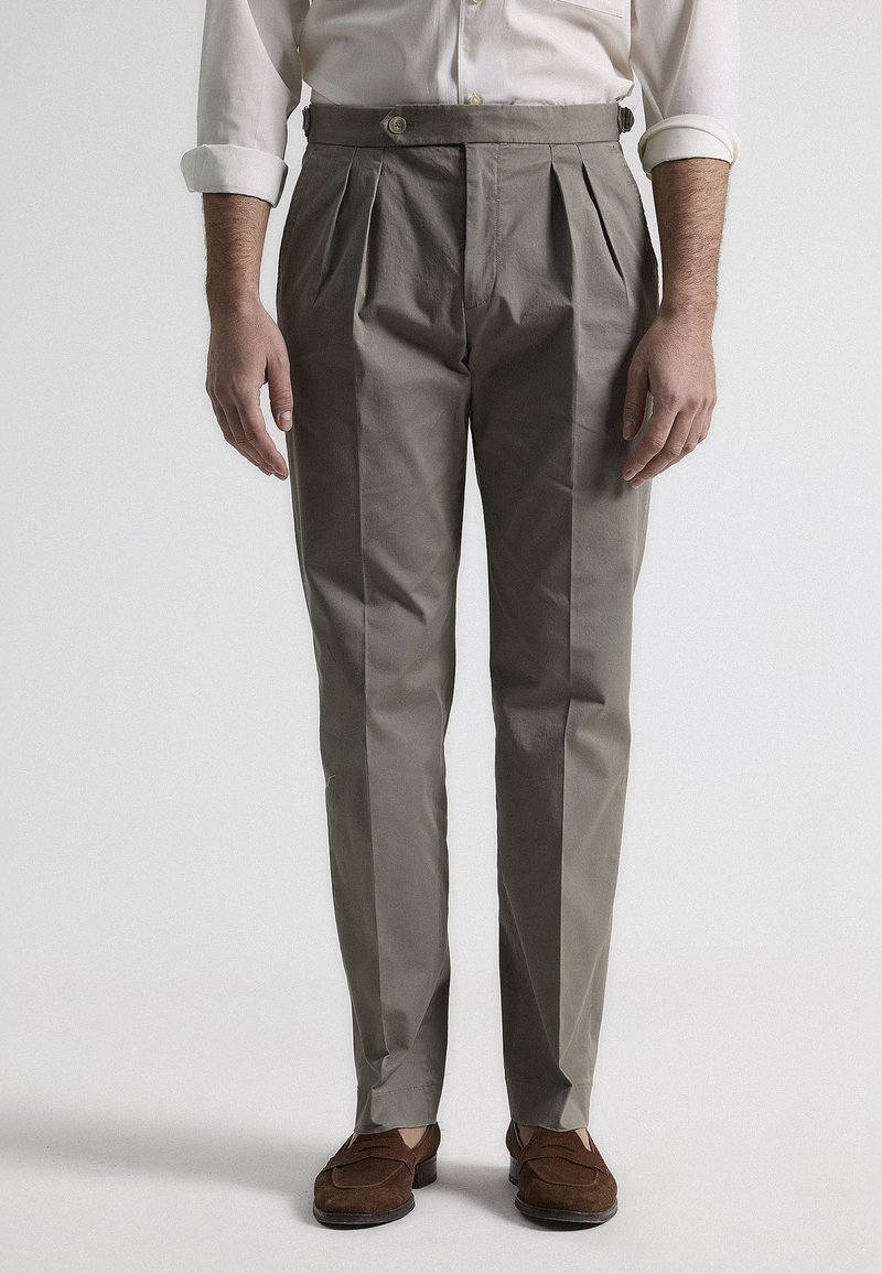 Pantalones de tejido khaki con pliegues y un corte ajustado, que cuentan con una cintura con botones, una textura suave y piernas rectas. Acentuados con zapatos de ante marrón.