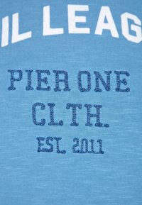 Camiseta de algodón de color azul claro con texto bordado: "PIER ONE CLTH. EST. 2011" en azul más oscuro, con letras grandes en blanco en la parte superior.