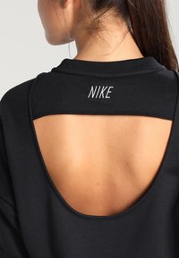 Czarna sportowa bluzka z szerokim, otwartym plecami. Logo "NIKE" wyszyte na biało przy dekolcie. Miękki, teksturowany materiał.