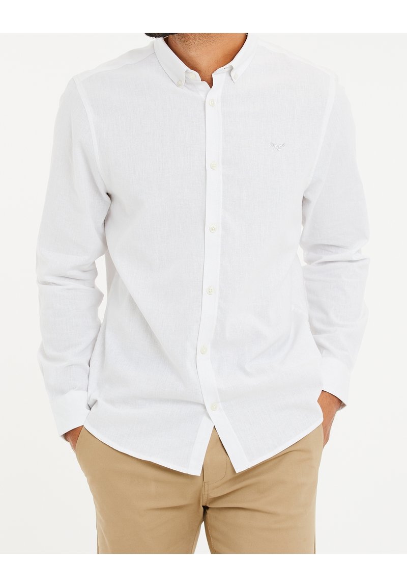 Homme portant une chemise blanche à manches longues avec col et un pantalon beige, les mains dans les poches, sur un fond uni.