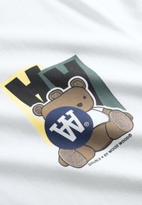 Brun teddybjørn, der holder en blå cirkel med hvide "W"-bogstaver, siddende foran gule og grønne klodser med sorte "W"-bogstaver på hvidt stof.