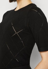 Svart kortärmad stickad topp med diamantmönstrade semitransparenta sektioner, bärs av en person som visas från halsen till midjan.