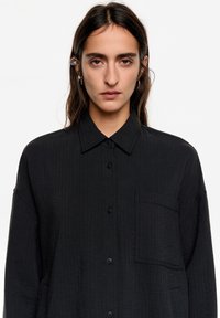 Camisa negra de manga larga con cierre de botones, cuello puntiagudo y un solo bolsillo. La tela texturada presenta un sutil patrón acanalado.