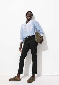 Blouse rayée bleu clair avec des détails brodés, jean noir ajusté et sabots en daim marron. Le mannequin porte un sac kaki.