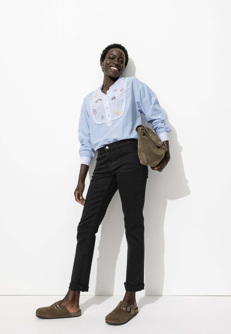 Blouse rayée bleu clair avec des détails brodés, jean noir ajusté et sabots en daim marron. Le mannequin porte un sac kaki.