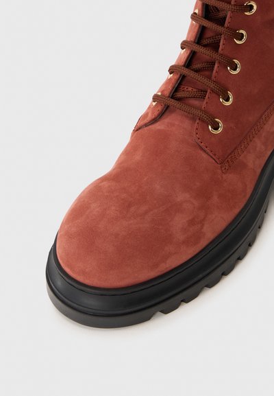 Brown suede ankle boot with a rounded toe, black rubber sole, and gold-tone eyelets for laces. Features textured surface and sturdy construction.