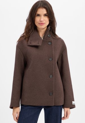 Woman wearing a brown buttoned wool coat with an asymmetrical collar and blue jeans against a white background.