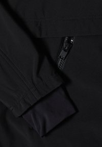 Gros plan d'une manche de veste noire avec un poignet élastique et une poche zippée noire partiellement visible.