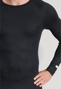 Haut thermique noir à manches longues, fabriqué en tissu ajusté et lisse, avec un col rond, des manches raglan et une petite étiquette blanche au poignet.